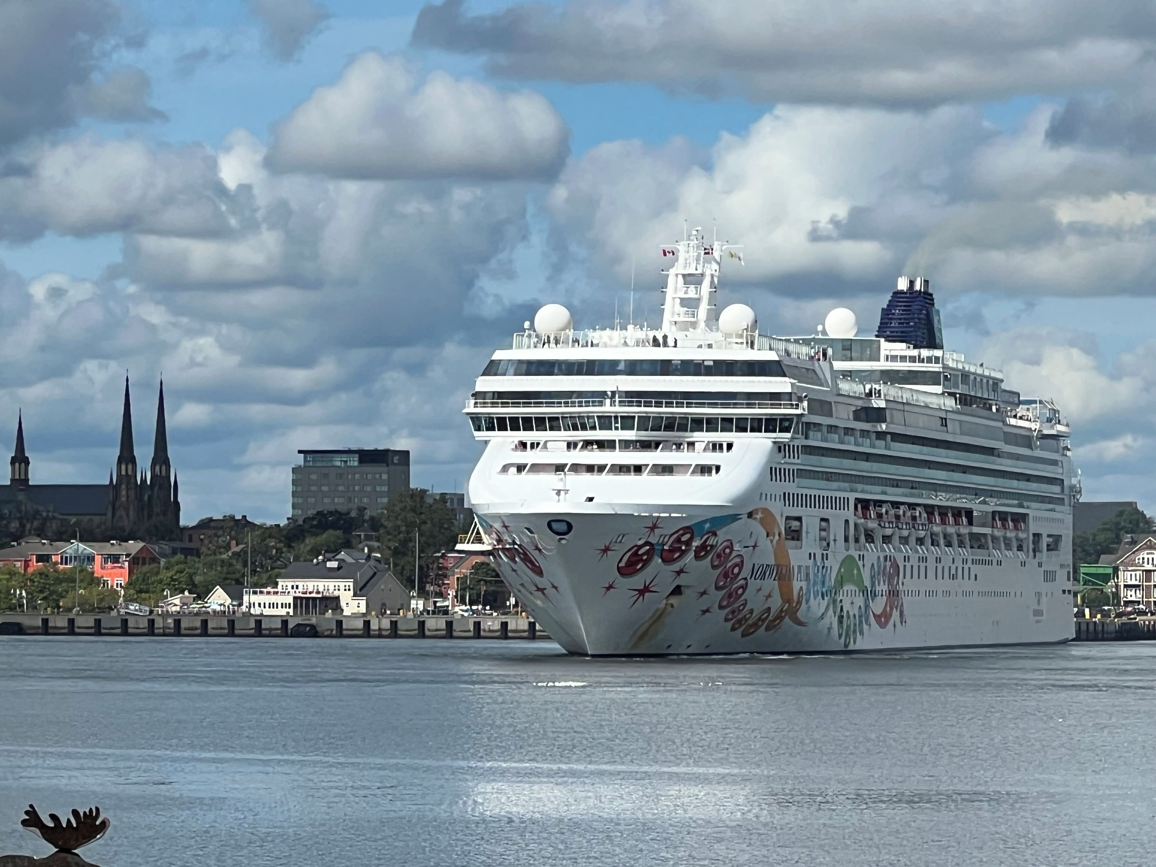 Et stort krydstogtskib ligger ved kaj i en havn, med en katedral og moderne bygninger i baggrunden.
