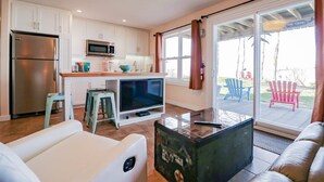 Una cocina moderna con armarios blancos, un refrigerador de acero inoxidable, un microondas y un televisor sobre un mueble. Hay una puerta corrediza de vidrio que da a un espacio exterior con sillas.