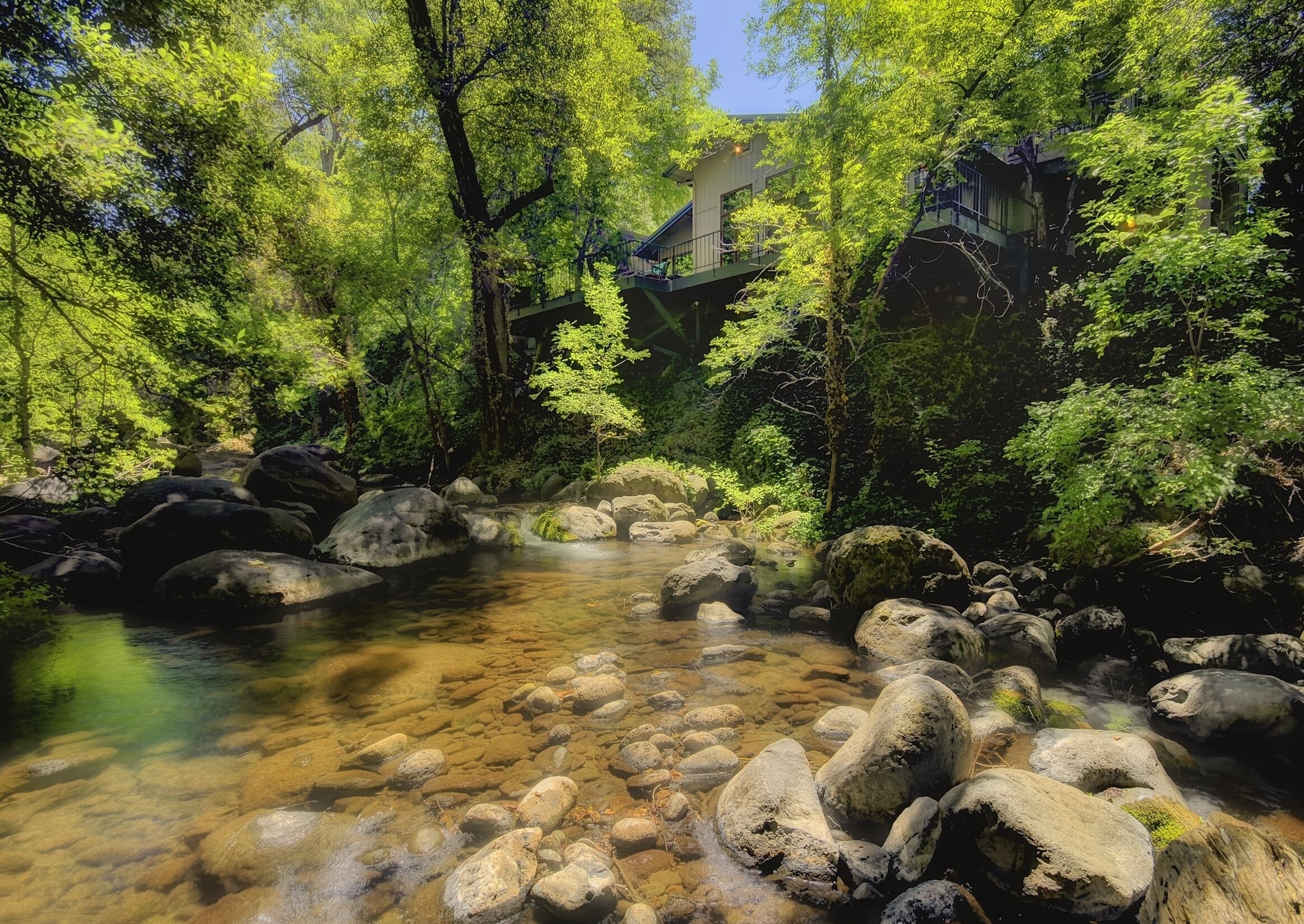 A clear stream with rocks, surrounded by lush green trees and a bridge in the background.