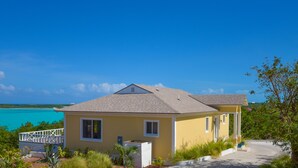 A two-story yellow house with a balcony, surrounded by greenery and a view of the ocean.