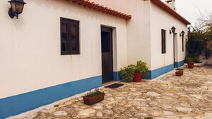 Ein Gebäude mit weißer Fassade, blauen Akzenten und einem Ziegeldach. Blumentöpfe und eine Tür sind zu sehen.