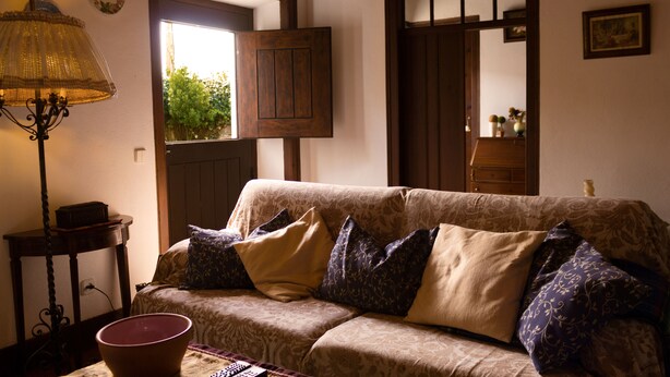Ein gemütliches Wohnzimmer mit einer gemusterten Couch, einer Lampe und Blick durch eine offene Tür.