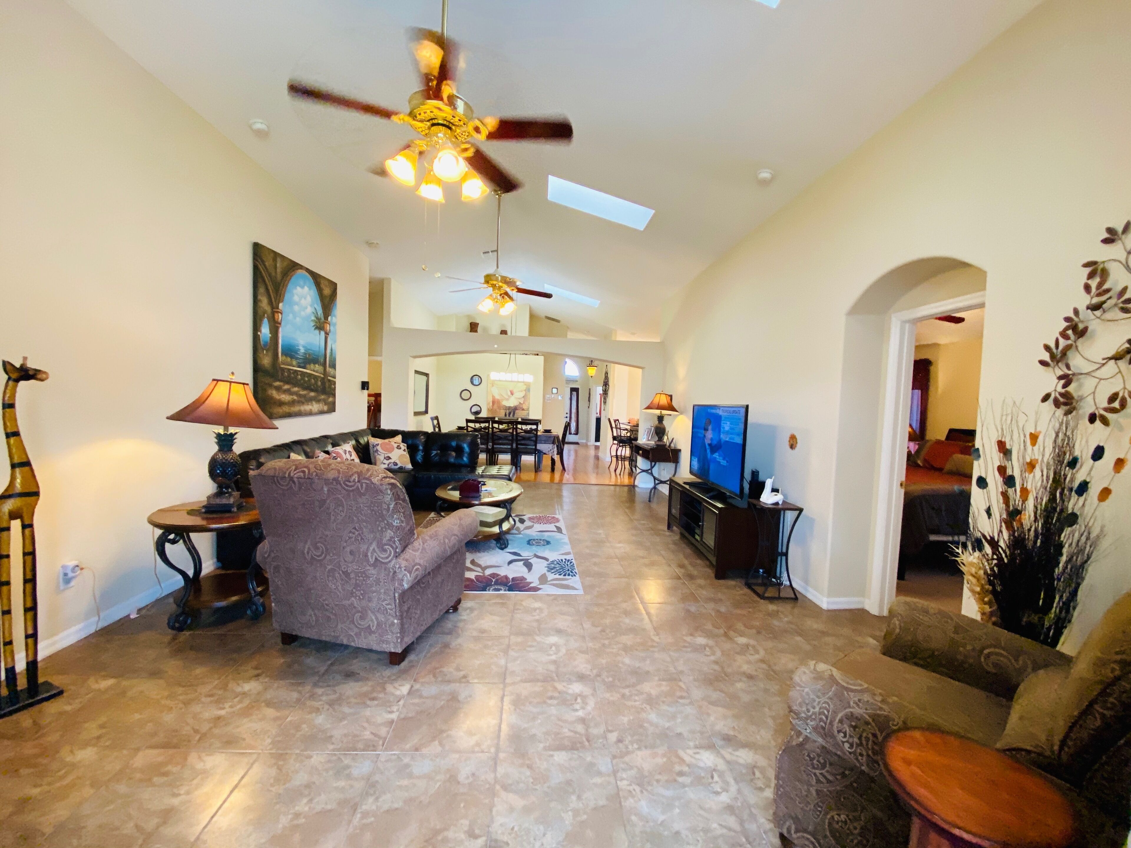 A spacious living area with a sofa, armchairs, a coffee table, and a television. There is a ceiling fan, a dining area in the background, and a decorative plant.