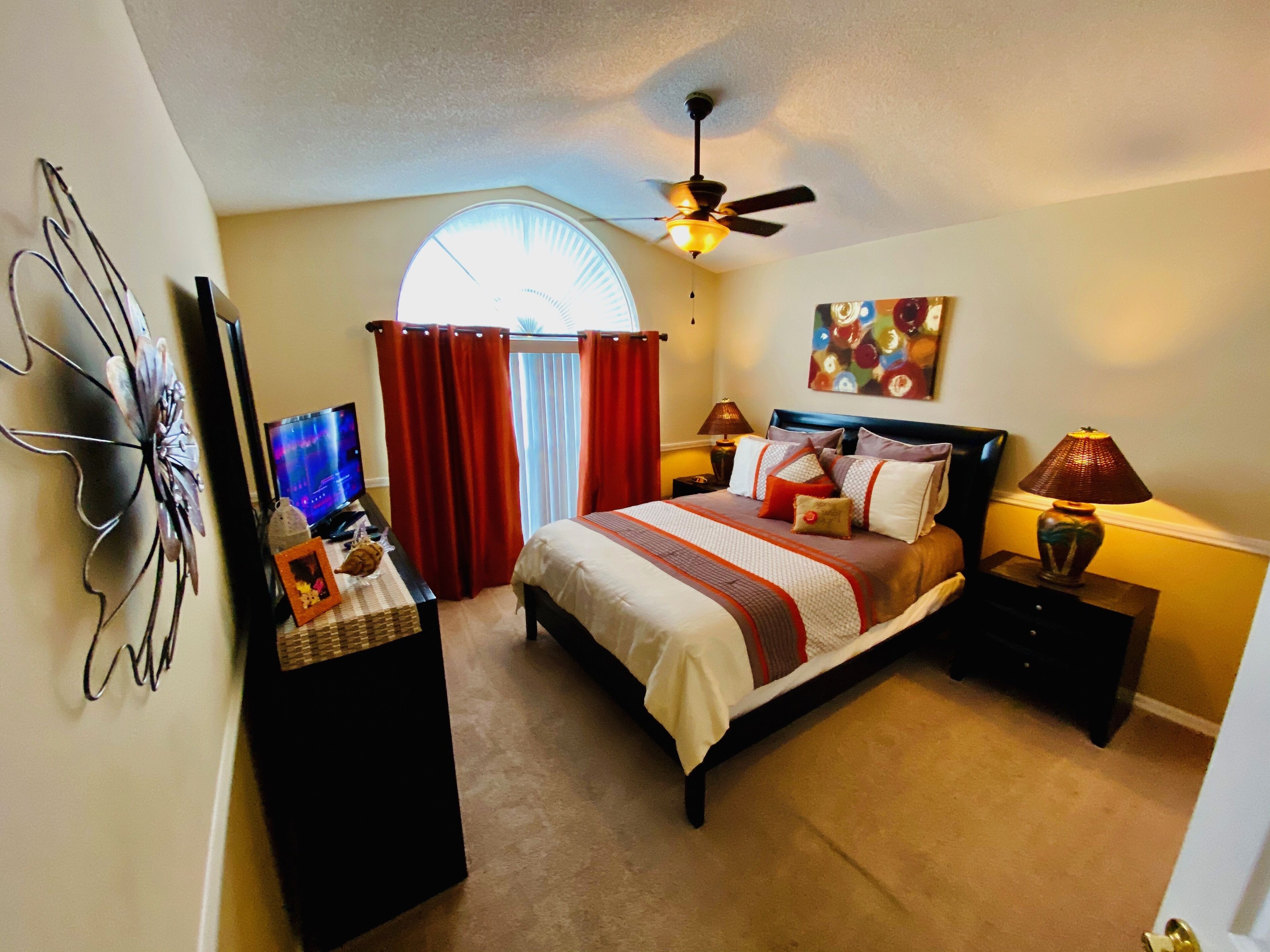 A bedroom with a bed, a ceiling fan, a TV, and a painting on the wall.