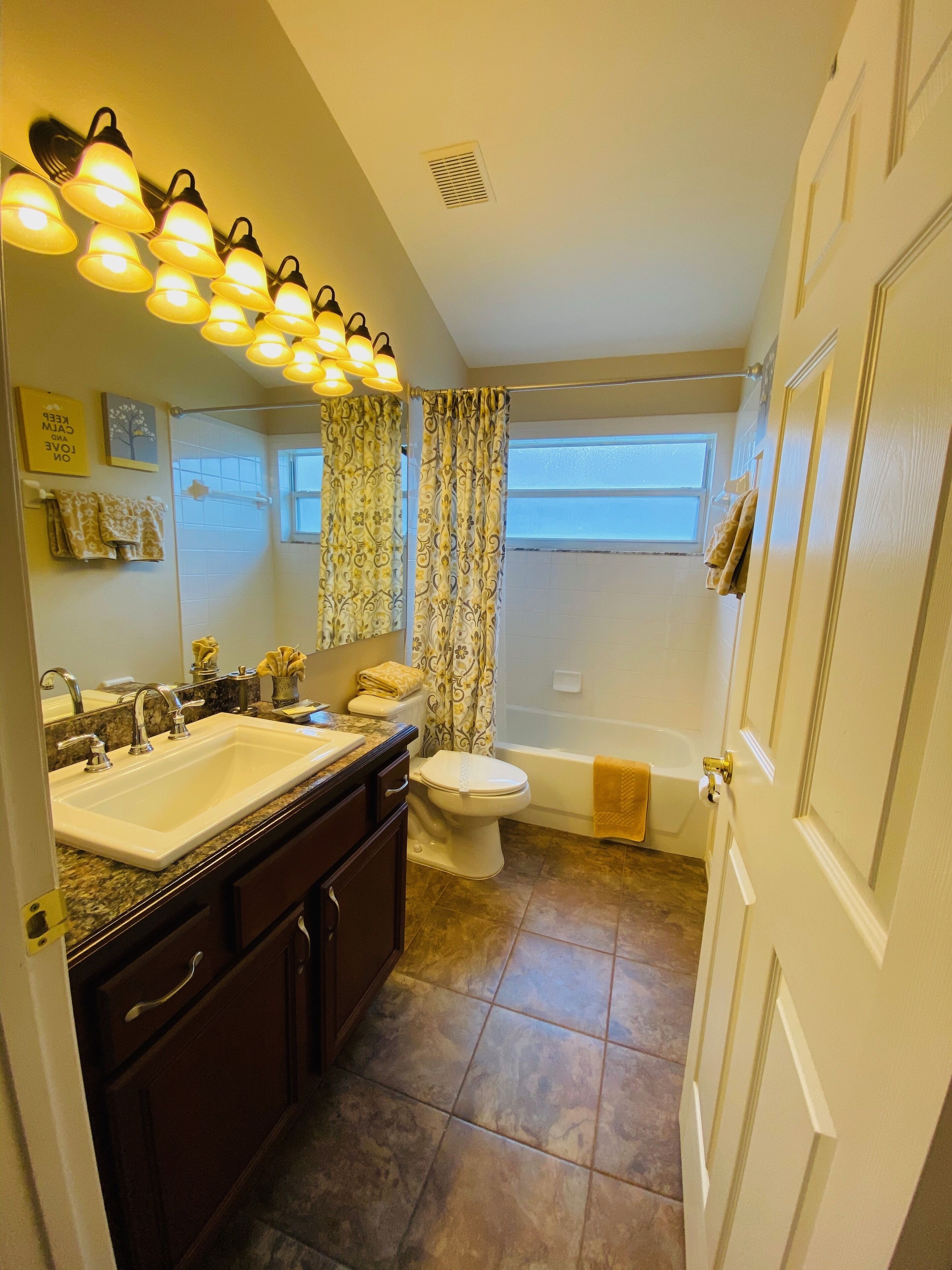 Une salle de bain  équipée d’un lavabo, de toilettes et d’une baignoire, avec des rideaux à motifs et un miroir agrémenté de lumières au-dessus.