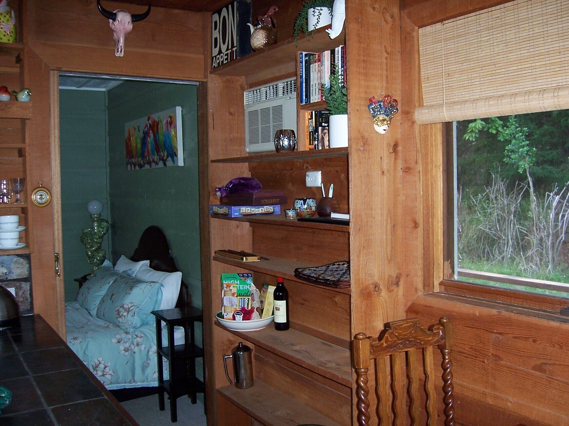 A cozy room with a bed, wooden shelves with books and decorative items, a window with a view of trees, and a door leading to another room.