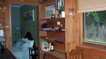A cozy room with a bed, wooden shelves with books and decorative items, a window with a view of trees, and a door leading to another room.