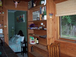 A cozy room with a bed, wooden shelves with books and decorative items, a window with a view of trees, and a door leading to another room.