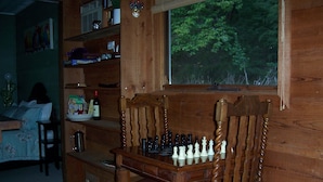 A wooden interior with a chessboard on a table, a window with a view of trees, and a bookshelf with various items. - The Huntsman Cabin (Warburton)