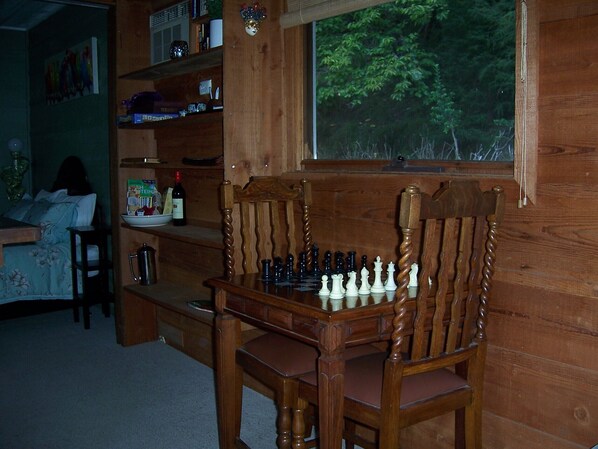 A wooden interior with a chessboard on a table, a window with a view of trees, and a bookshelf with various items.