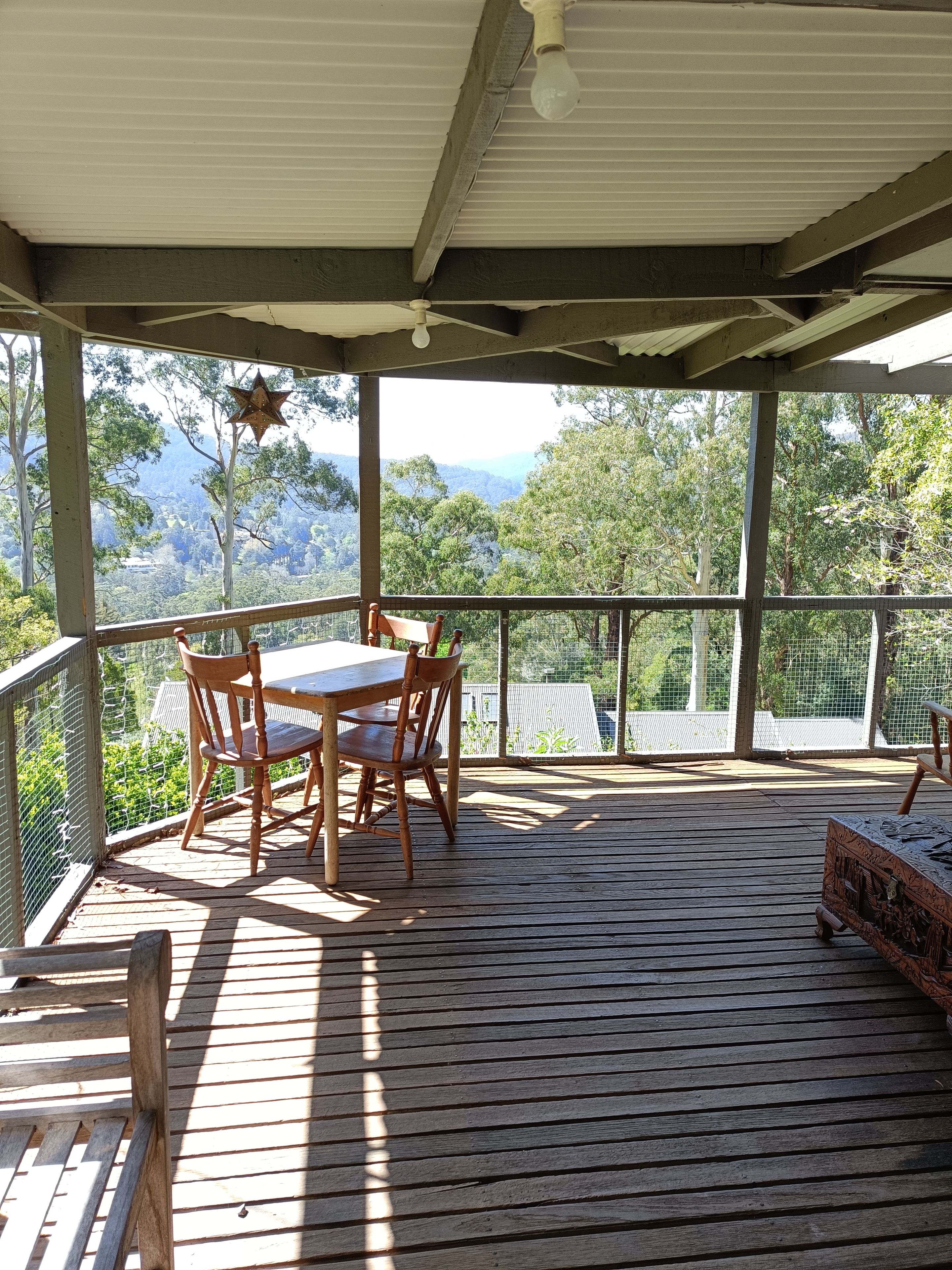 Un espacio al aire libre techado con una mesa y sillas, vista a árboles y montañas, y una terraza de madera.