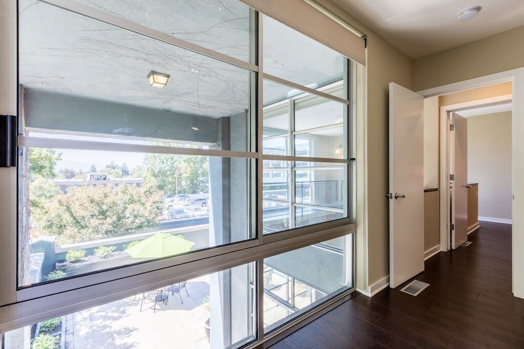 A room with large windows, a wooden floor, and a door leading to another room.