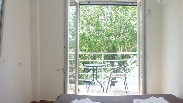 Un dormitorio con cama, un balcón con mesa y sillas, y una puerta abierta al exterior.
