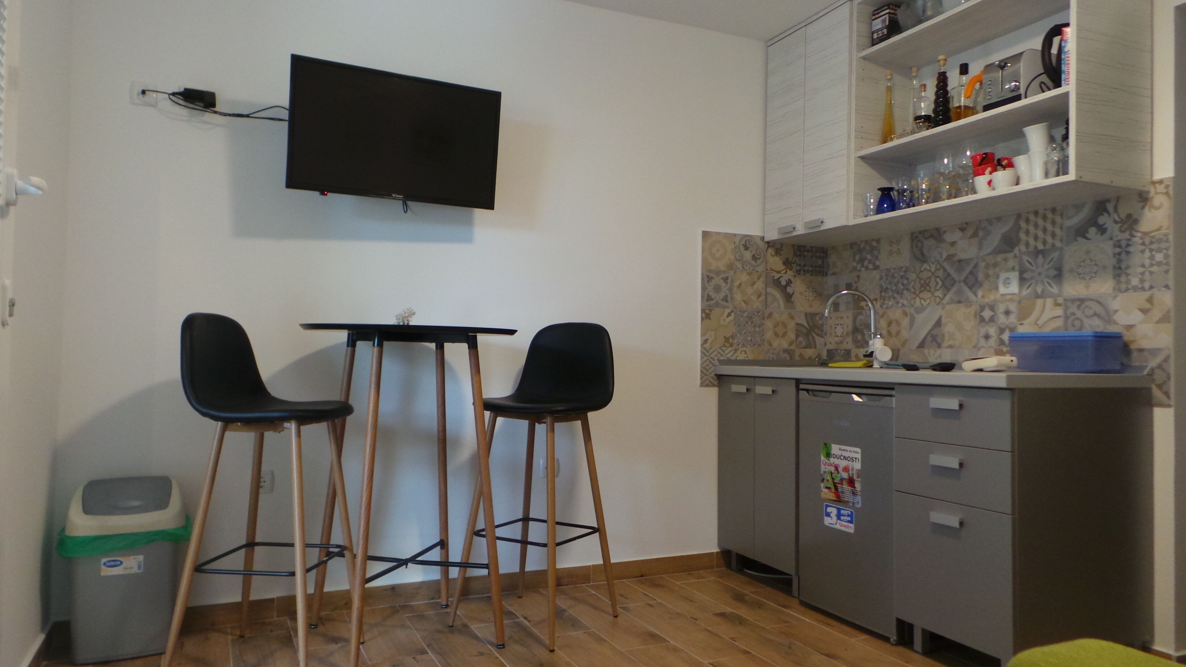 Une petite cuisine avec un téléviseur fixé au mur, deux chaises noires, une table en bois, un évier et un plan de travail carrelé.
