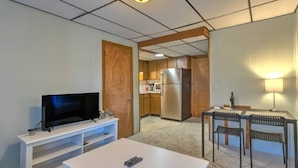 Um espaço de estar compacto com televisão em um suporte branco, uma mesa pequena com um controle remoto, uma cozinha compacta com geladeira e micro-ondas, e uma área de jantar com duas cadeiras e uma mesa.