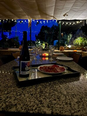 Een terras met een eettafel gedekt voor twee personen, met een fles Riesling, een bord met parmaham en een kaars als decoratie.