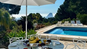 Ein Essbereich am Pool mit einem Tisch für zwei Personen, einem weißen Sonnenschirm und Liegestühlen.