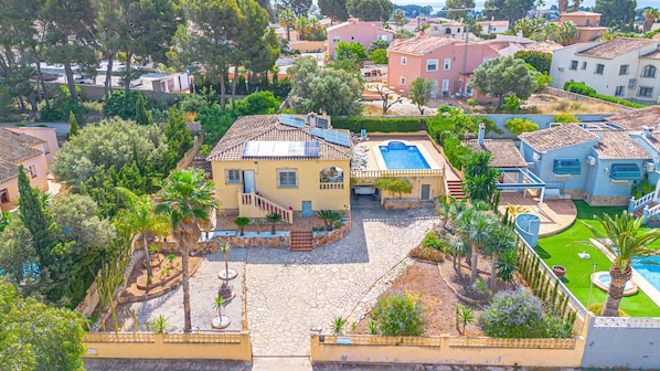 A residential area with a large house featuring a swimming pool, a patio, and a garden with palm trees.