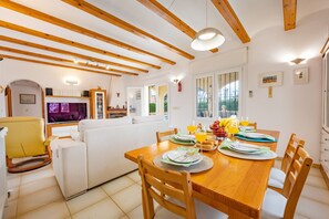 A spacious living area with a wooden dining table set for four, surrounded by wooden chairs. A white sofa and a yellow rocking chair are visible in the background.