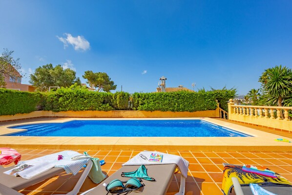 A swimming pool with lounge chairs and towels, surrounded by greenery and a clear sky.