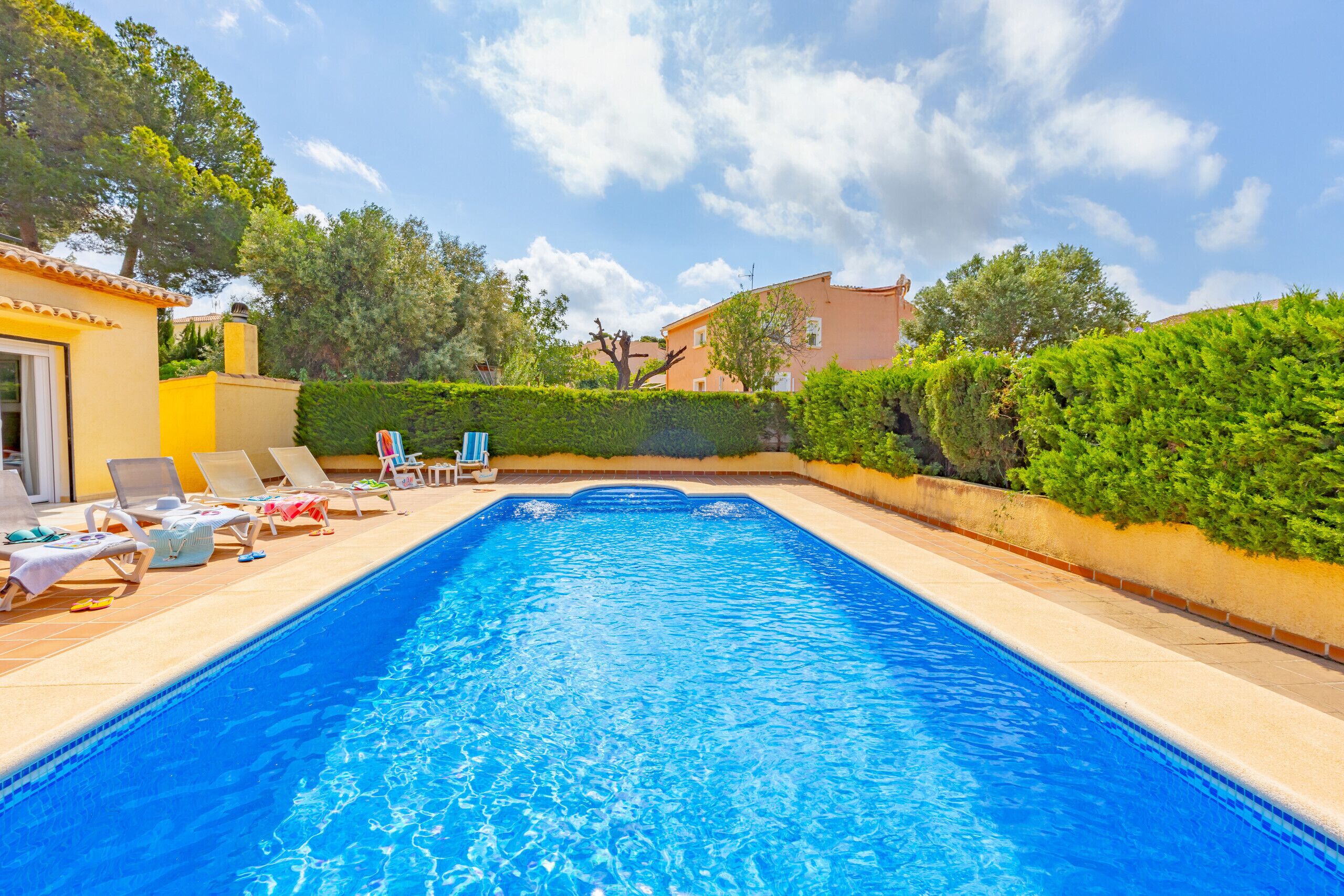 A rectangular swimming pool with clear blue water, surrounded by a tiled deck. There are lounge chairs and a small table with a towel.