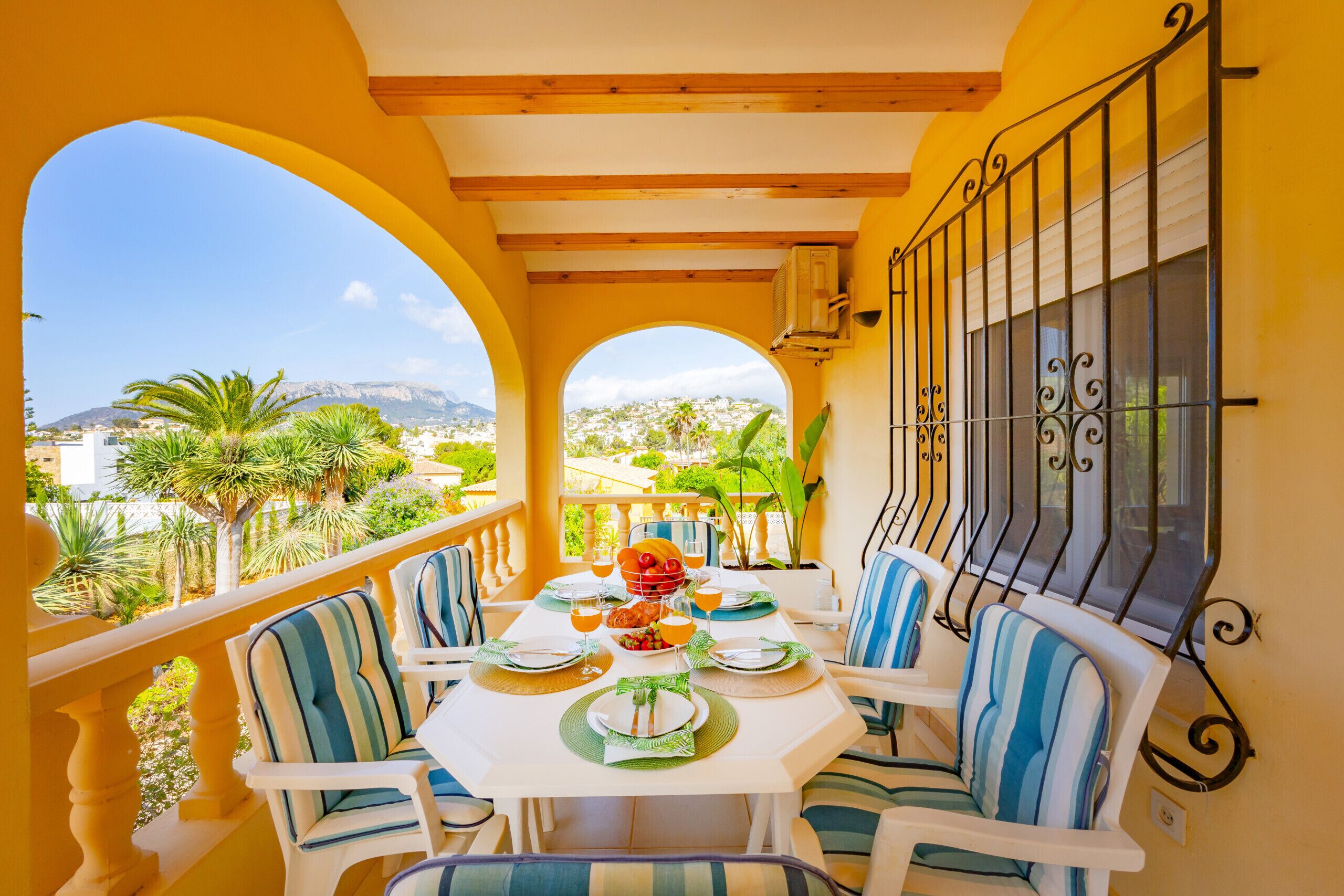 A dining area with a table set for four, surrounded by chairs with striped cushions, a view of the landscape, and a balcony railing.