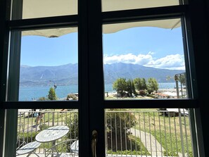 Une vue à travers une fenêtre ornée d’une rambarde en métal, avec une table et des chaises, donnant sur un lac et des montagnes.