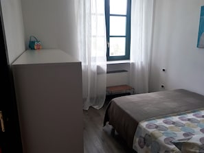 A bedroom with a bed, a small table, a window with curtains, and a wall-mounted shelf. - Campione Giulia Exclusive Lakefront (Campione del Garda)