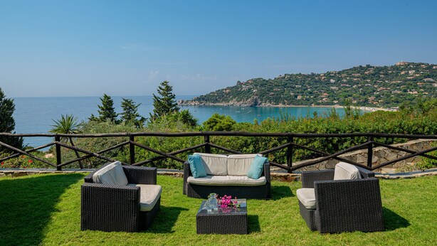 Une terrasse dotée de meubles en osier, d’une table en verre et offrant une vue sur la mer et les collines.