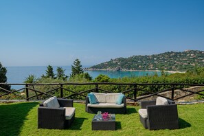 A patio with wicker furniture, a glass table, and a view of the sea and hills.