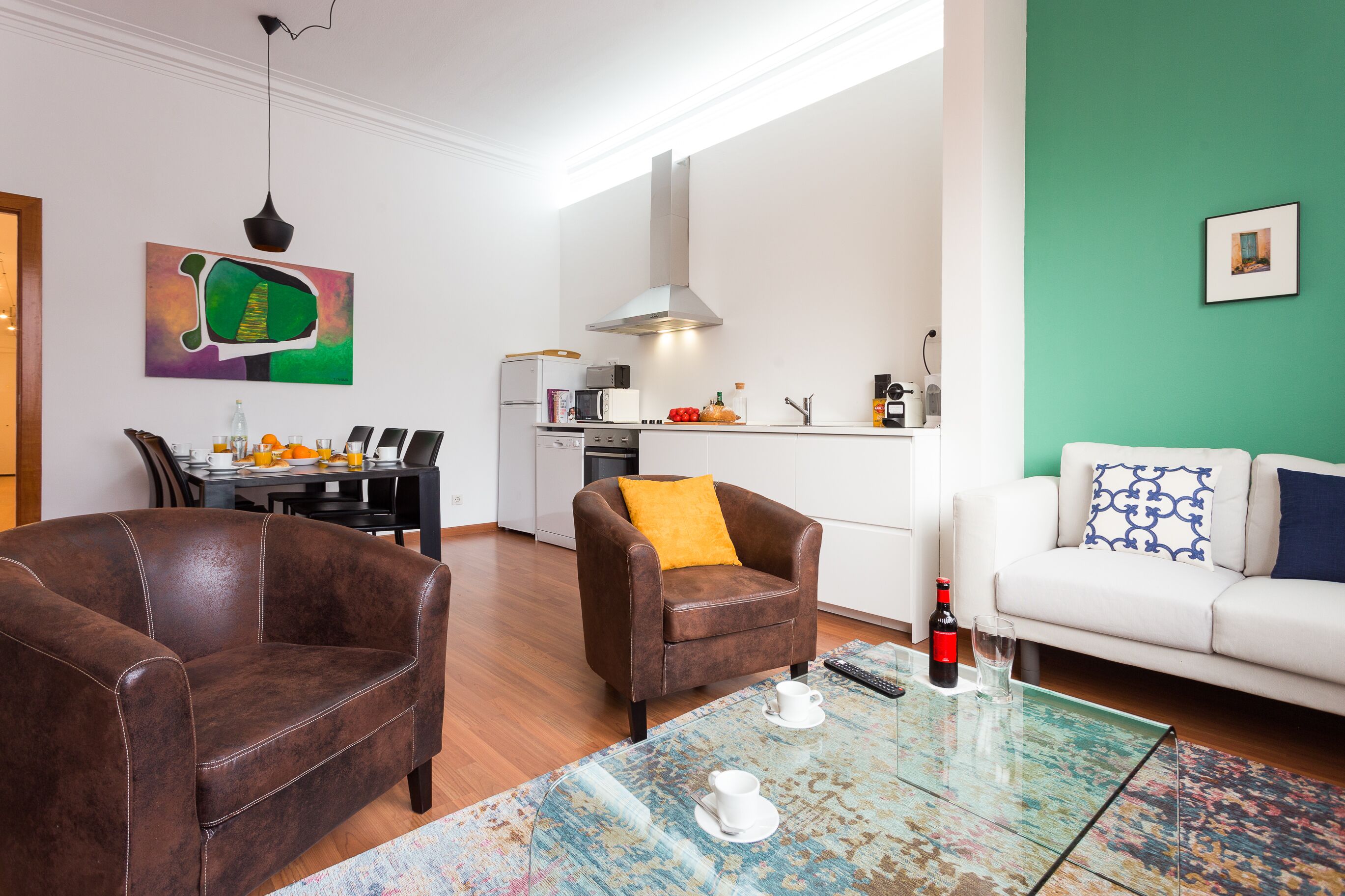 A modern living room with leather sofas, a glass coffee table, and a kitchen in the background.