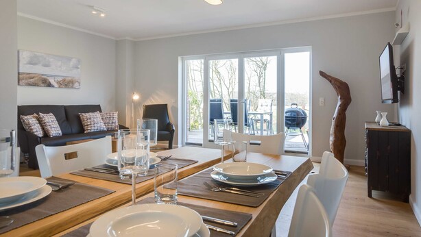 A dining area with a wooden table set for four, a sofa with patterned cushions, and a large window with a view of the outdoors. - Fehmarn - Fehmarn in List on Sylt (List)