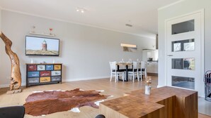 A modern living room with a wooden coffee table, a leather rug, a television mounted on the wall, and a dining area with white chairs and a glass table. - Fehmarn - Fehmarn in List on Sylt (List)