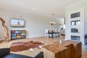A modern living room with a wooden coffee table, a leather rug, a television mounted on the wall, and a dining area with white chairs and a glass table.