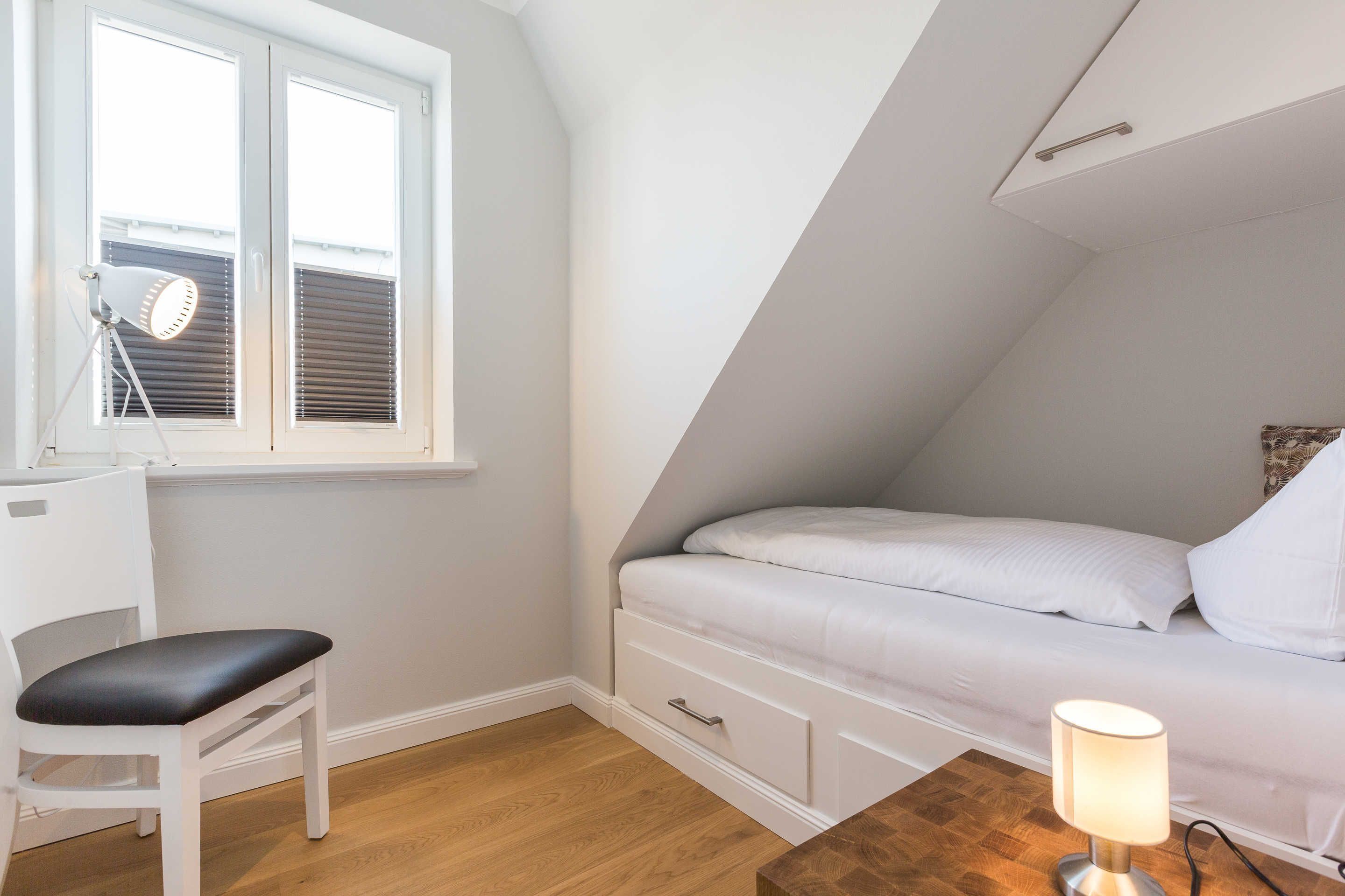 Ett sovrum med en säng, ett skrivbord med en stol, en lampa och ett fönster med persienner.