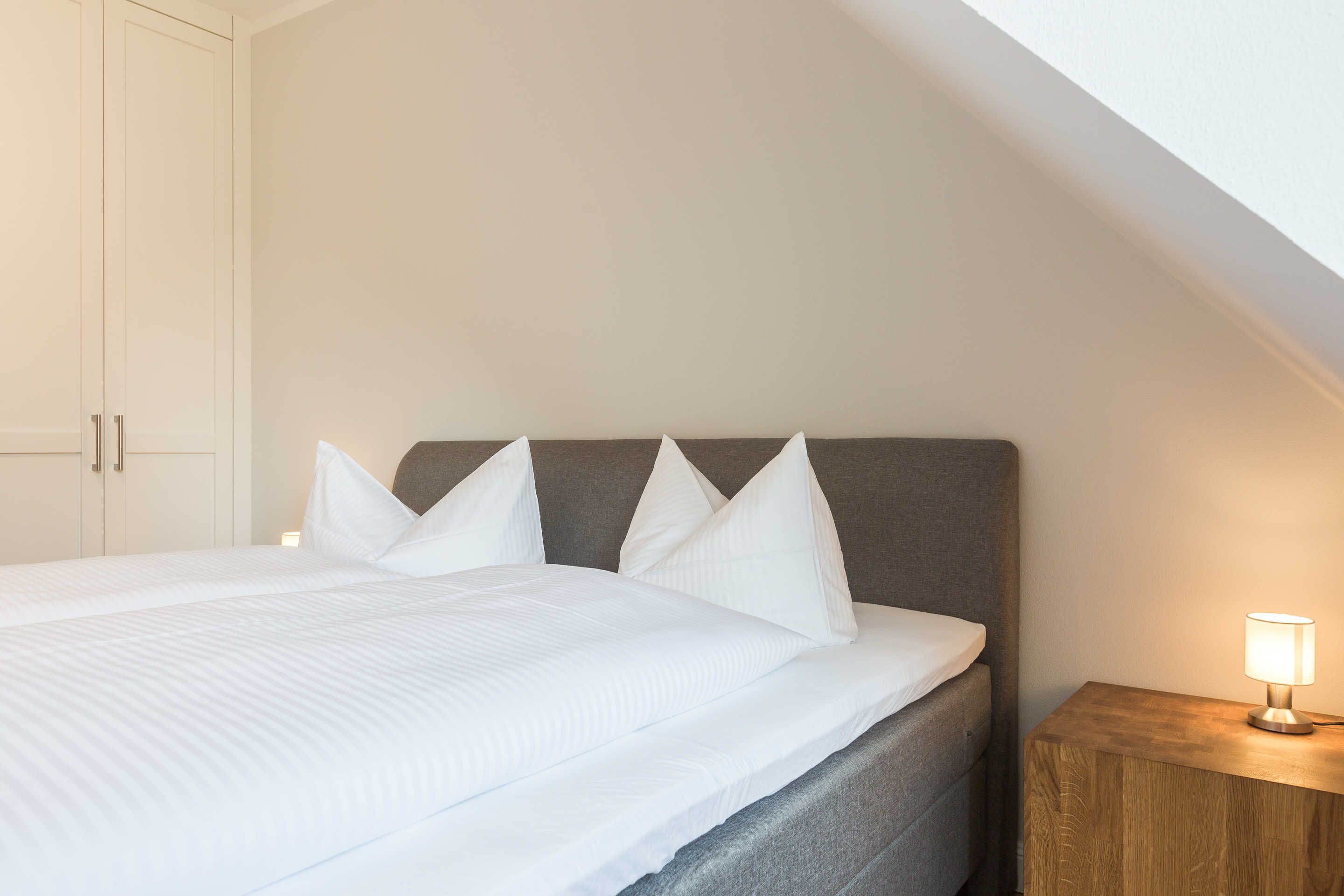 A neatly made bed with white linens and pillows, a wooden nightstand with a lamp, and a white wardrobe.