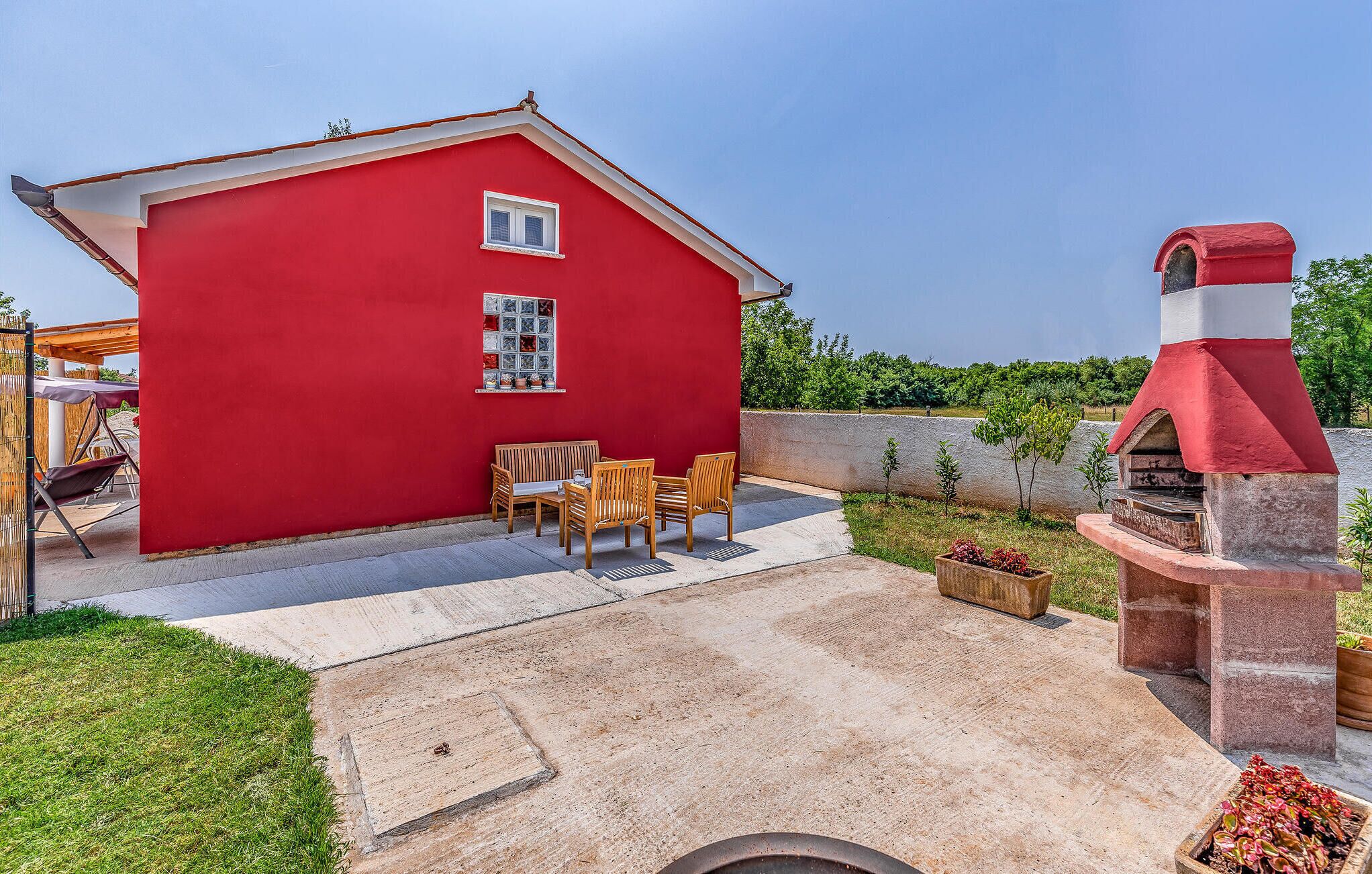 A red house with a small patio, a wooden table and chairs, and a stone barbecue grill.