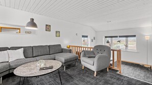 En moderne stue med en grå sofa, et rundt sofabord og en grå lænestol. Der er gelænder nær et vindue med udsigt til det fri.