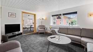 En moderne stue med en grå sofa, et rundt sofabord, et fjernsyn og et spiseområde med stole og bord.