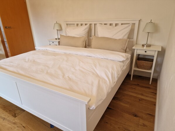 Una cama blanca con cabecera de madera, dos mesitas de noche con lámparas, y una cesta de mimbre sobre una de ellas.