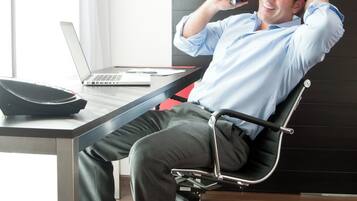 Un hombre sentado en una silla de oficina moderna frente a un escritorio con una computadora portátil, un teléfono y un altavoz negro.