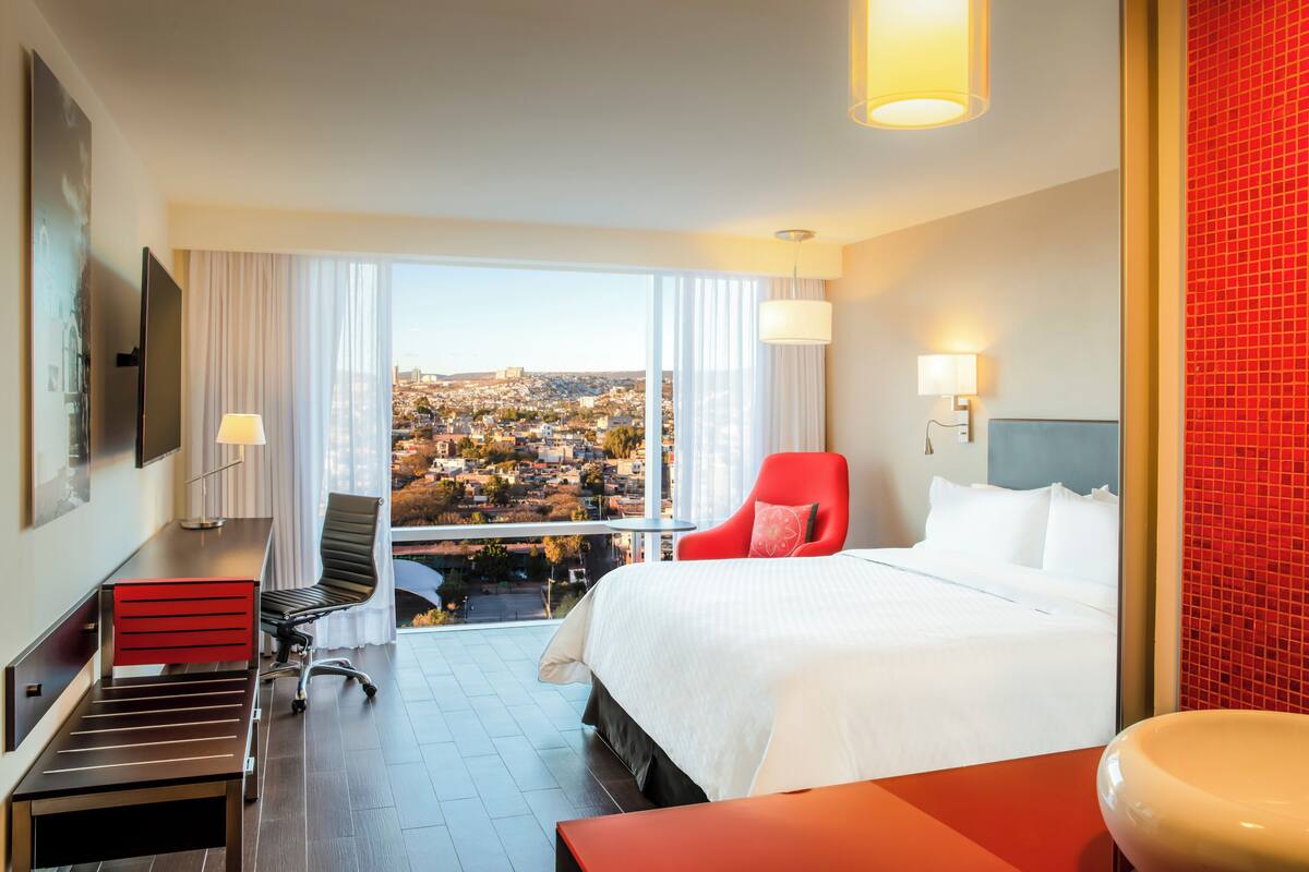 A hotel room with a large bed, a red chair, a desk, and a view of a cityscape through large windows.