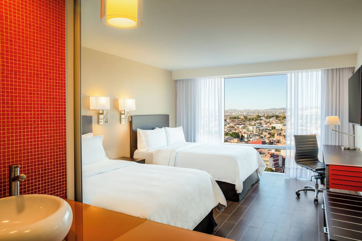 A hotel room with two beds, a desk, a chair, a large window with a city view, and a red-tiled wall.