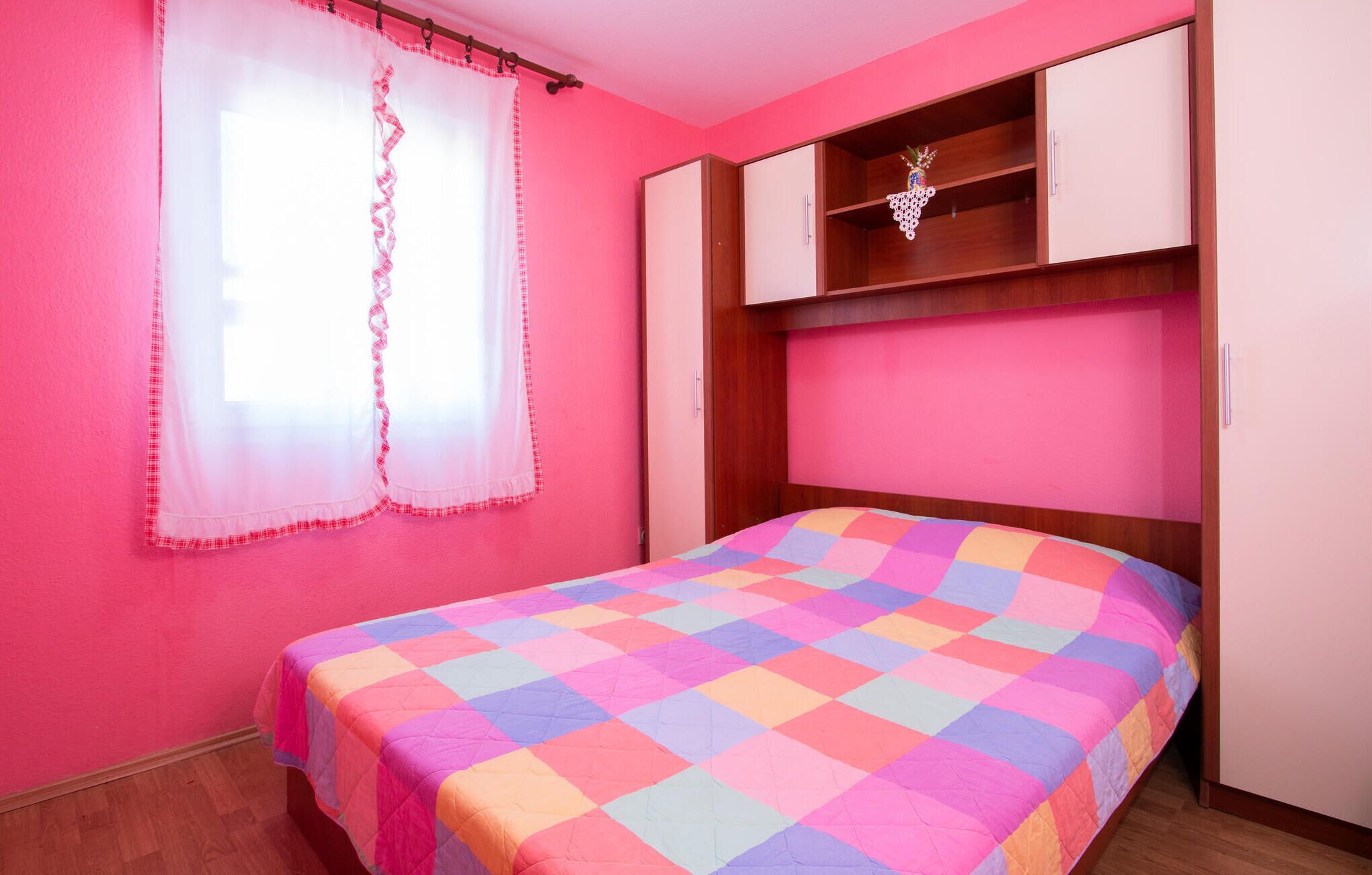 A bedroom with pink walls, a bed with a colorful quilt, a wooden wardrobe, and a window with sheer curtains.