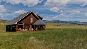 A wooden cabin with a large porch and outdoor seating, surrounded by a grassy field and mountains in the background. - Yellowstone Trail Lodge · Yellowstone National park (Emigrant)