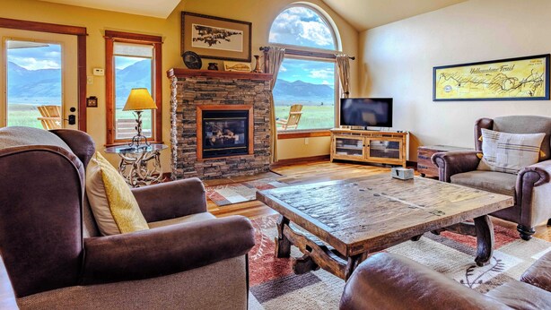 A cozy living room with a stone fireplace, leather sofas, a wooden coffee table, and a large window offering a view of mountains. - Yellowstone Trail Lodge · Yellowstone National park (Emigrant)