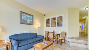Ein Wohnzimmer mit blauem Sofa, einem Holztisch und einem Essbereich mit Holzstühlen und einem Tisch. An der Wand hängt ein gerahmtes Gemälde und es gibt ein Fenster mit Fensterläden.