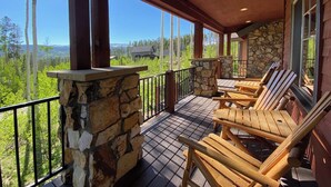 Una terraza de madera con sillas Adirondack, muros de piedra y grandes ventanales con vistas a un paisaje montañoso.