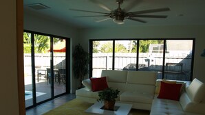 A spacious living room with a white sofa, a coffee table, and a ceiling fan. Large windows provide a view of the backyard.