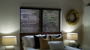 A bedroom with a bed, two bedside lamps, a window with blinds, and a wreath on the wall.